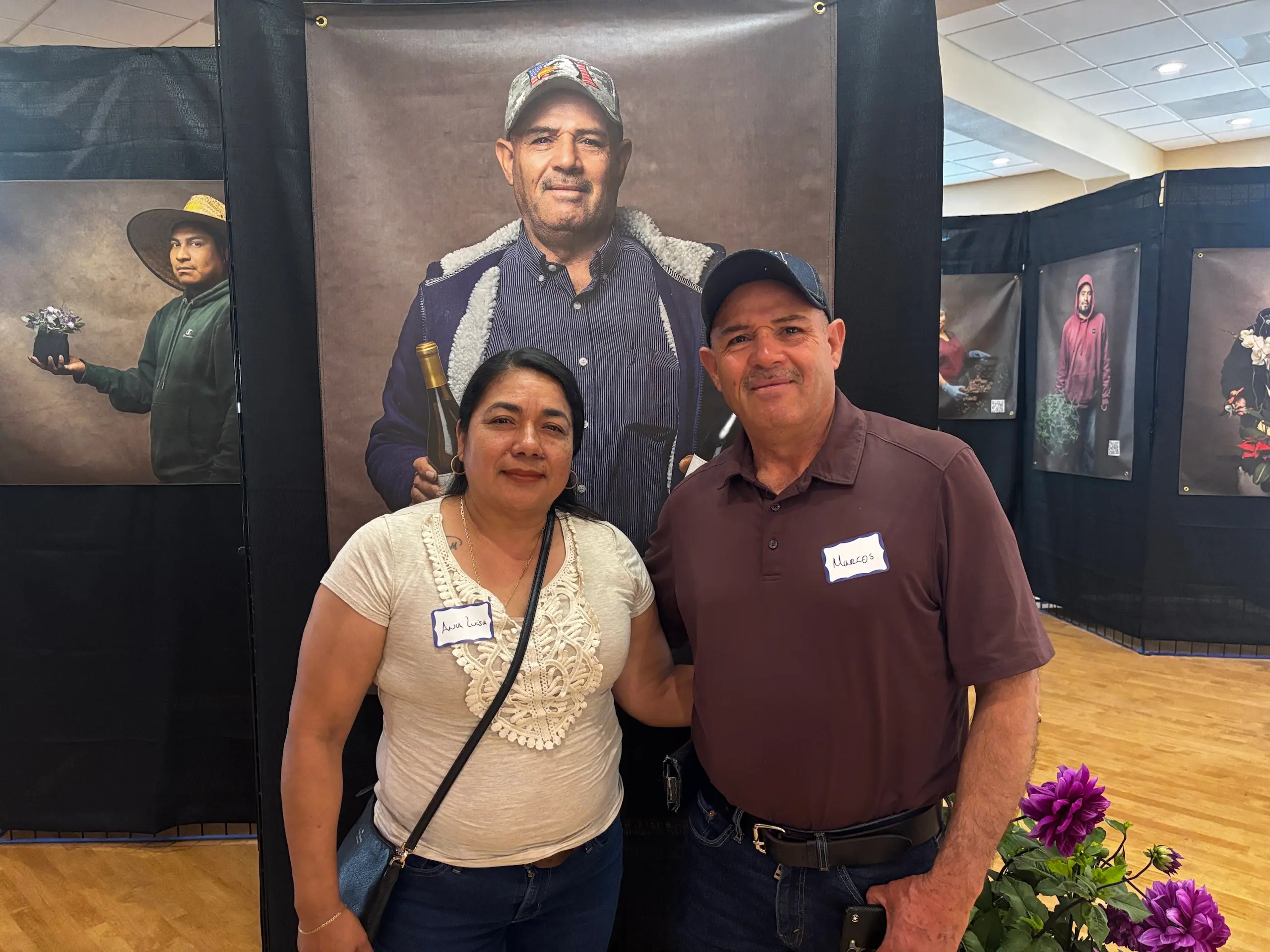 Marcos Jaimes y su esposa Ana Luisa delante del retrato de Marcos en la exposición. Marcos trabaja en un viñedo y en la foto exhibió con orgullo dos botellas de vino del lugar donde labora.