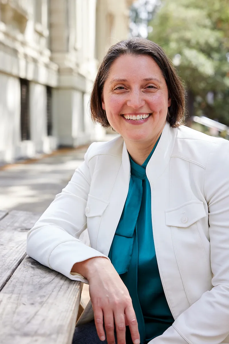 A person with short brown hair, wearing a white jacket over a teal blouse, smiles while leaning on a wooden bench outdoors.