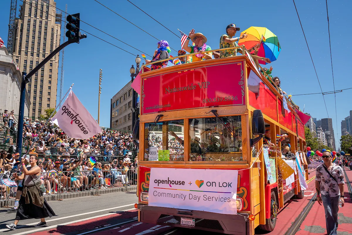 A vibrant parade features a colorful double-decker trolley adorned with rainbow flags and banners, carrying joyful participants as crowds cheer from the sidelines under a clear blue sky.