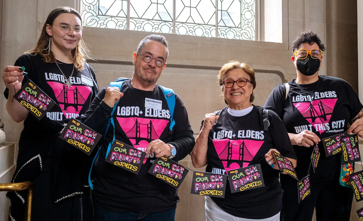 Four individuals wearing matching "LGBTQ+ Elders" shirts and holding "Our City Stories" cards stand together indoors near a decorative window.