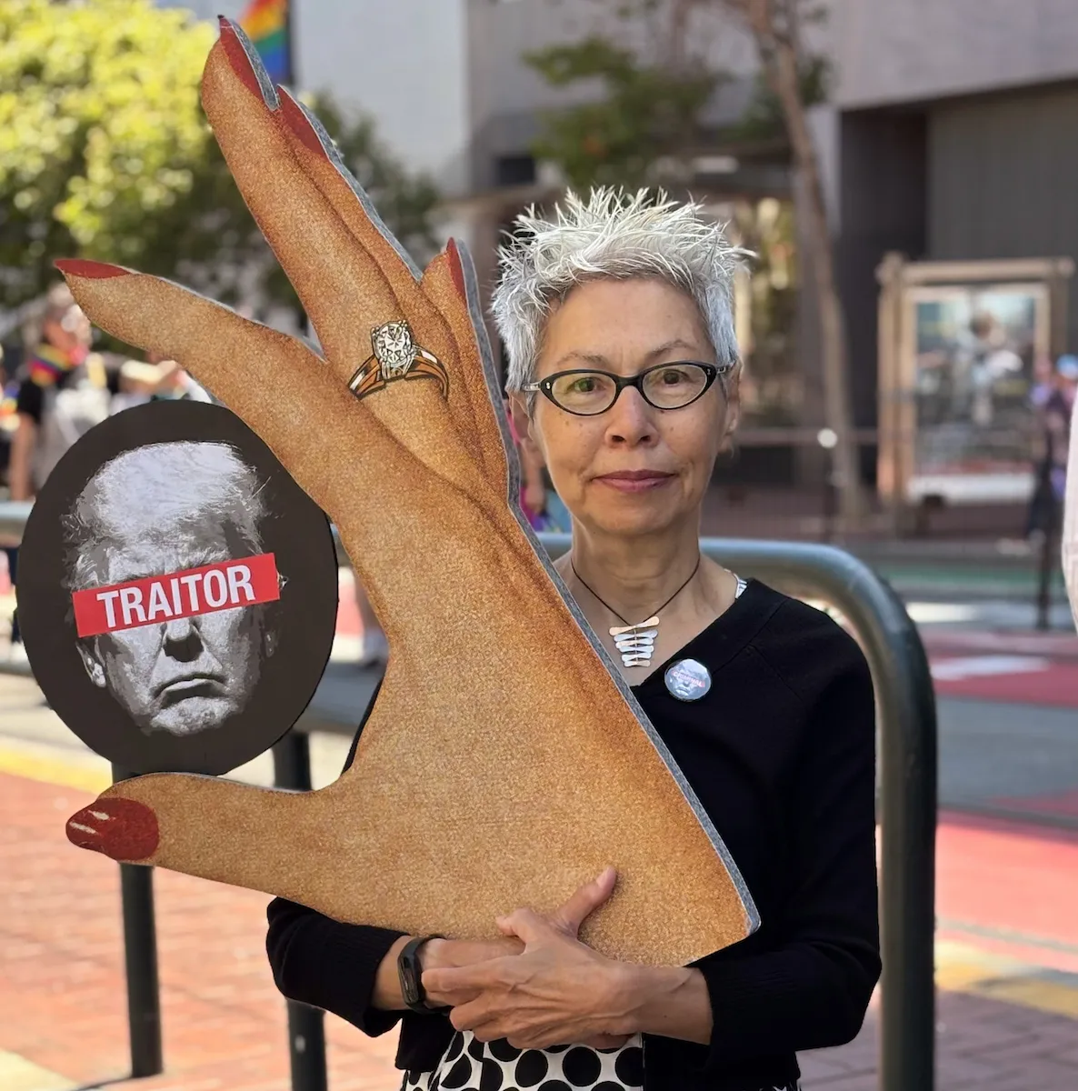 A person with short white hair and glasses holds a large cardboard hand prop with a circular cutout displaying a portrait labeled "TRAITOR" and a ring on the finger.
