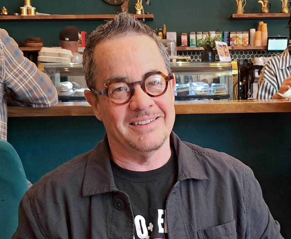 A person with short gray hair and round glasses smiles while seated in a café, wearing a dark jacket over a black t-shirt.
