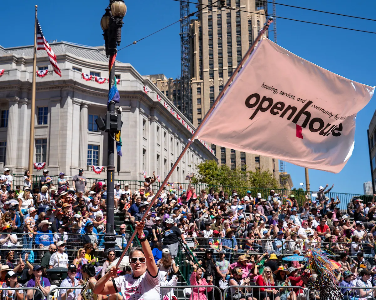 A person waves a large pink "openhouse" flag in front of a large crowd gathered in a city square under a clear blue sky.
